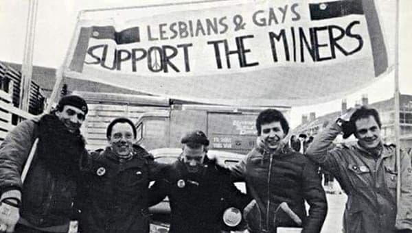 Five men stood in a row, arms over each other's shoulders, posing for the camera and holding a banner that says, ‘Lesbians & Gays Support the Miners’.