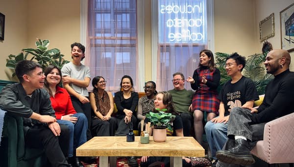11 people sit across three sofas facing each other in a brightly lit room, one is sat on the floor. They are smiling and laughing. A coffee table with plants is in front of them.