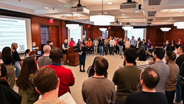 A large group of students standing in a semi-circle facing the front in a Harvard lecture room. Marshall Ganz is standing at the front behind a podium and Stephanie Wong is stood alongside speaking into a handheld microphone.
