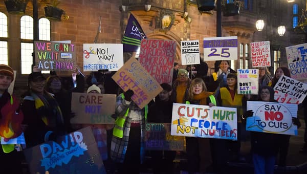 A group of people protesting, lit by street lights. They are holding placards with various slogans against council funding cuts.