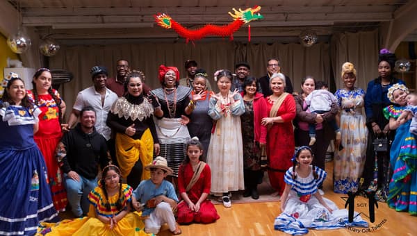 A diverse group of around 25 adults and children in traditional cultural dress from various backgrounds, posing together on stage at the Sanctuary Got Talents event, with a decorative Chinese dragon hanging above them.