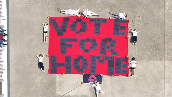 A birds eye view from high in the air showing a huge banner laid flat on concrete reading, 'Vote for home'. there are people lying on the floor around the banner.