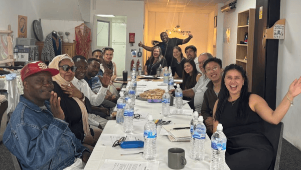A dozen people sitting around a long table, looking at the camera, smiling and waving.