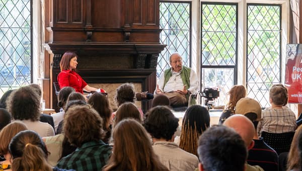 Stephanie Wong and Marshall Ganz are sat on chairs on a stage in front of an audience. Marshall Ganz is talking and gesturing towards the audience.