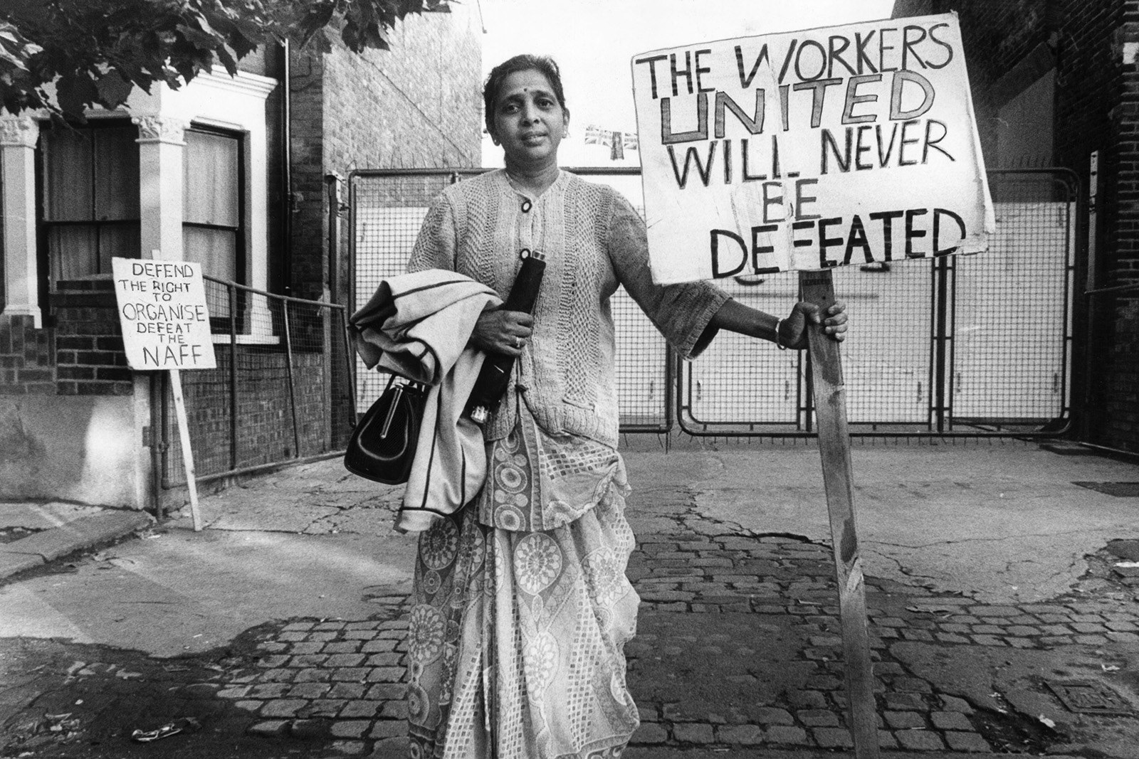 A middle-aged South-Asian woman stands smiling defiantly on a street. She wears traditional clothing and holds a sign saying 'The Workers United Will Never Be Defeated' - behind her another placard leans against the wall. It reads 'Defend the right to organise defeat the NAFF.' This image is black and white.