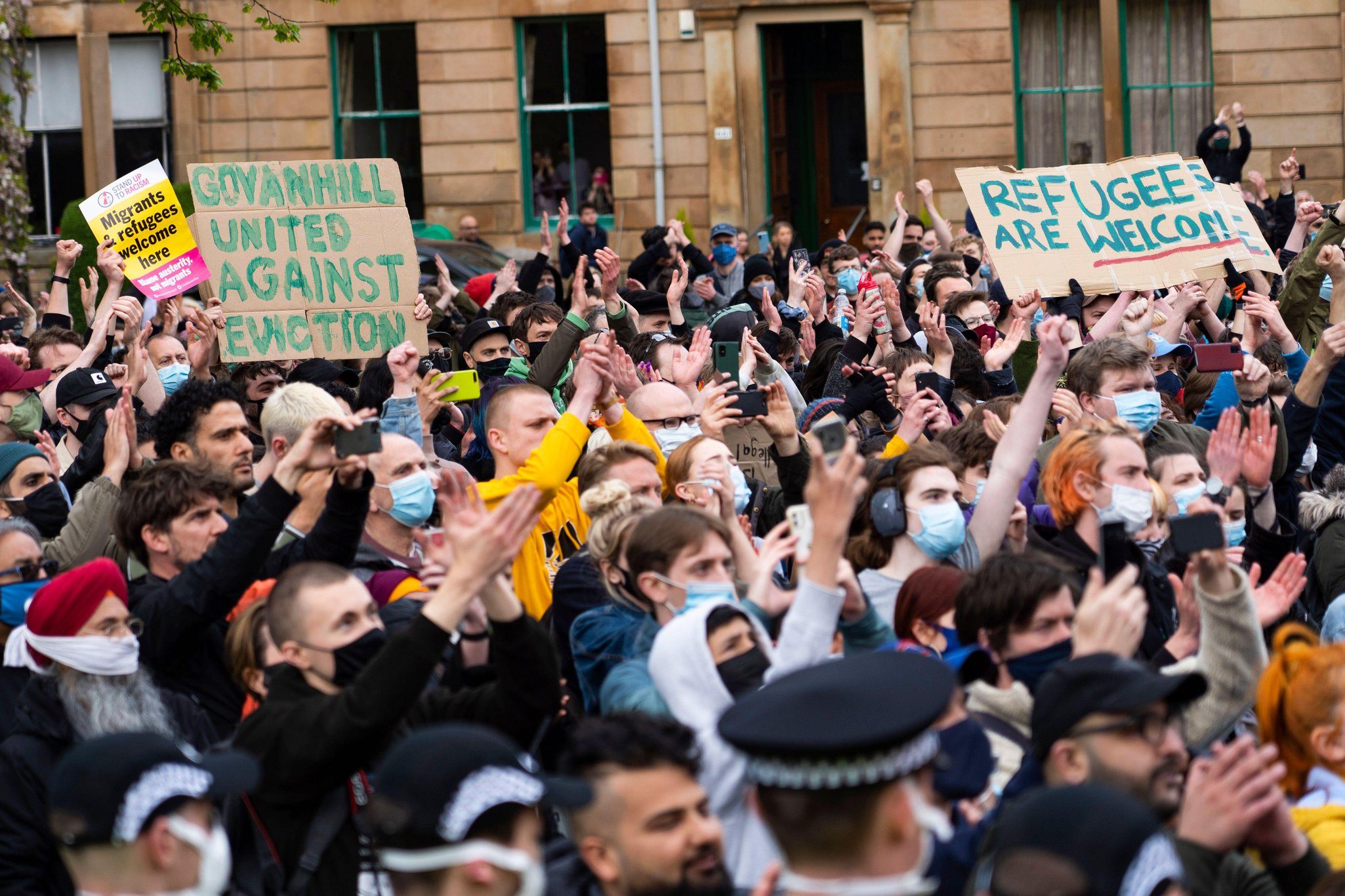 Protestors fill Kenmure Street. Some of them hold signs, these read: 'Refugees are welcome' 'Govanhill United Against Eviction' and 'Migrants and refugees welcome here'