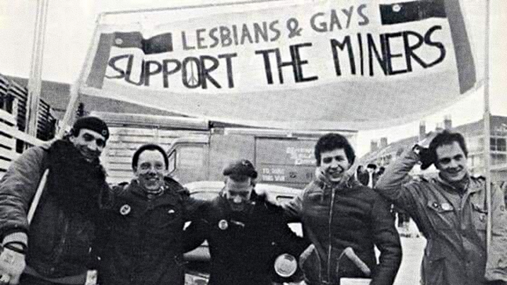 Five men stood in a row, arms over each other's shoulders, posing for the camera and holding a banner that says, ‘Lesbians & Gays Support the Miners’.