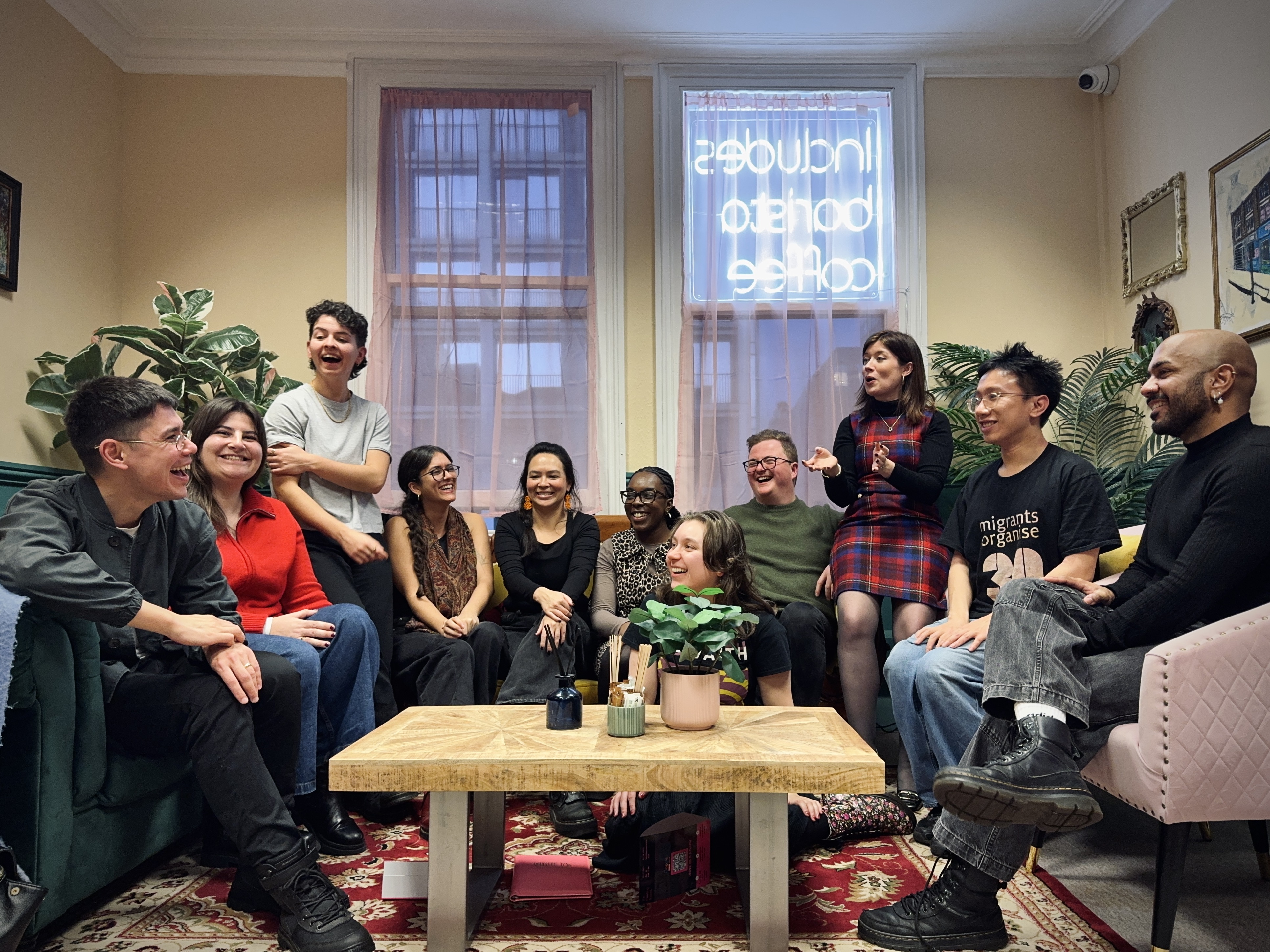 11 people sit across three sofas facing each other in a brightly lit room, one is sat on the floor. They are smiling and laughing. A coffee table with plants is in front of them.