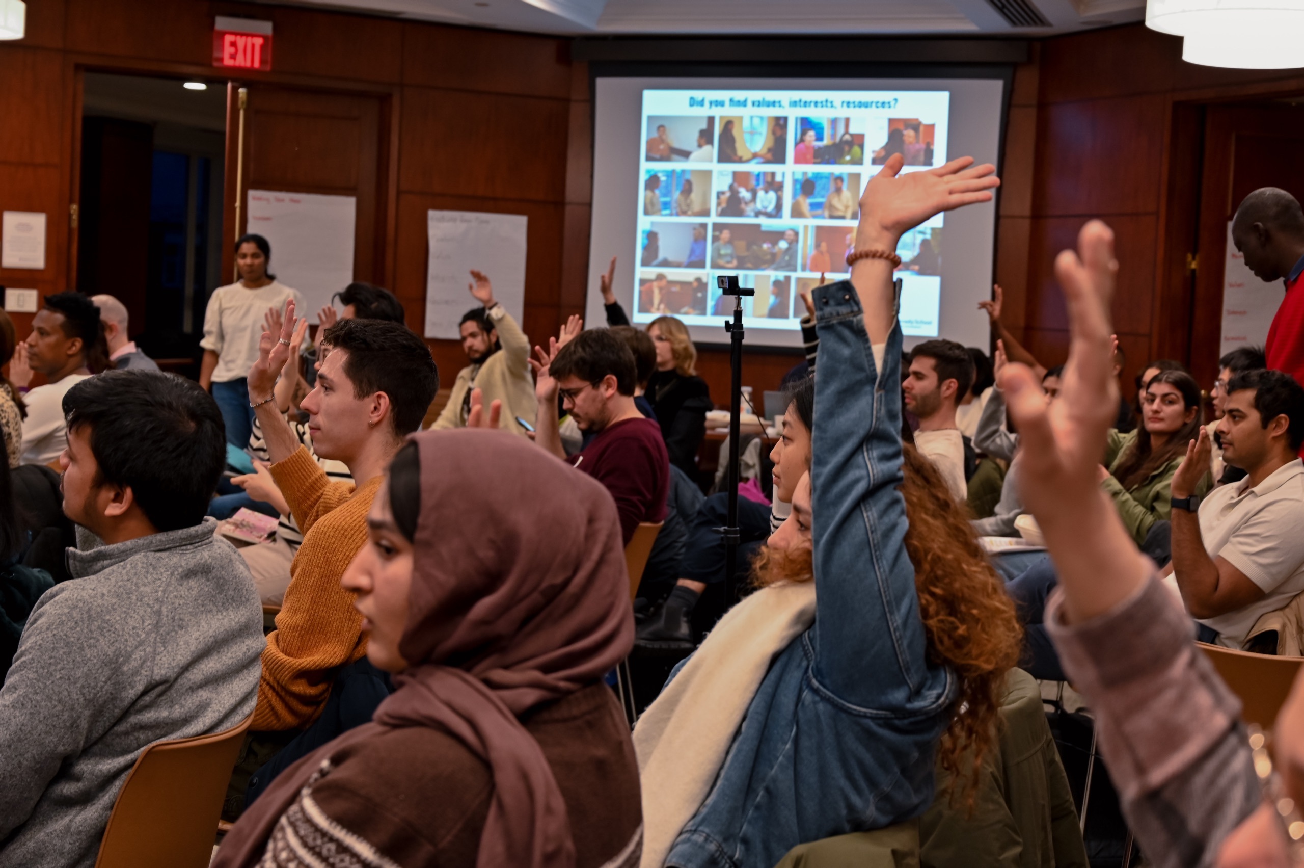 A large group of students sat facing in one direction in a Harvard lecture room. Many students have hands in the air.