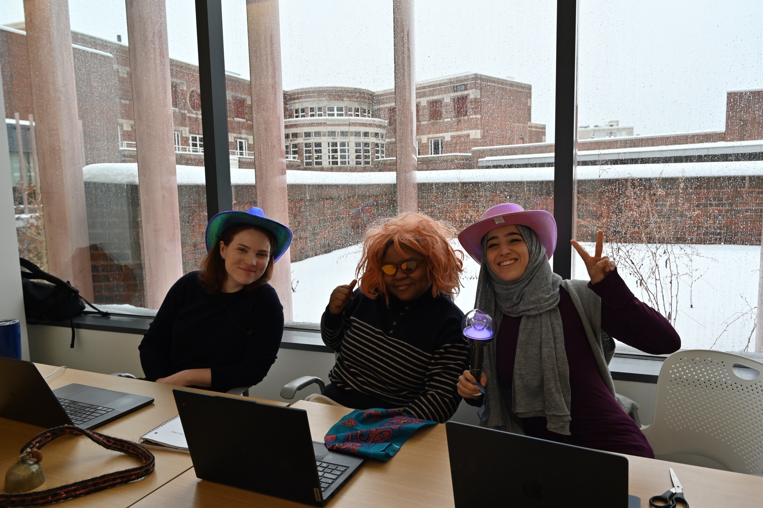 Three people at a desk posing for the camera wearing an assortment of colourful wigs, hats and glasses.
