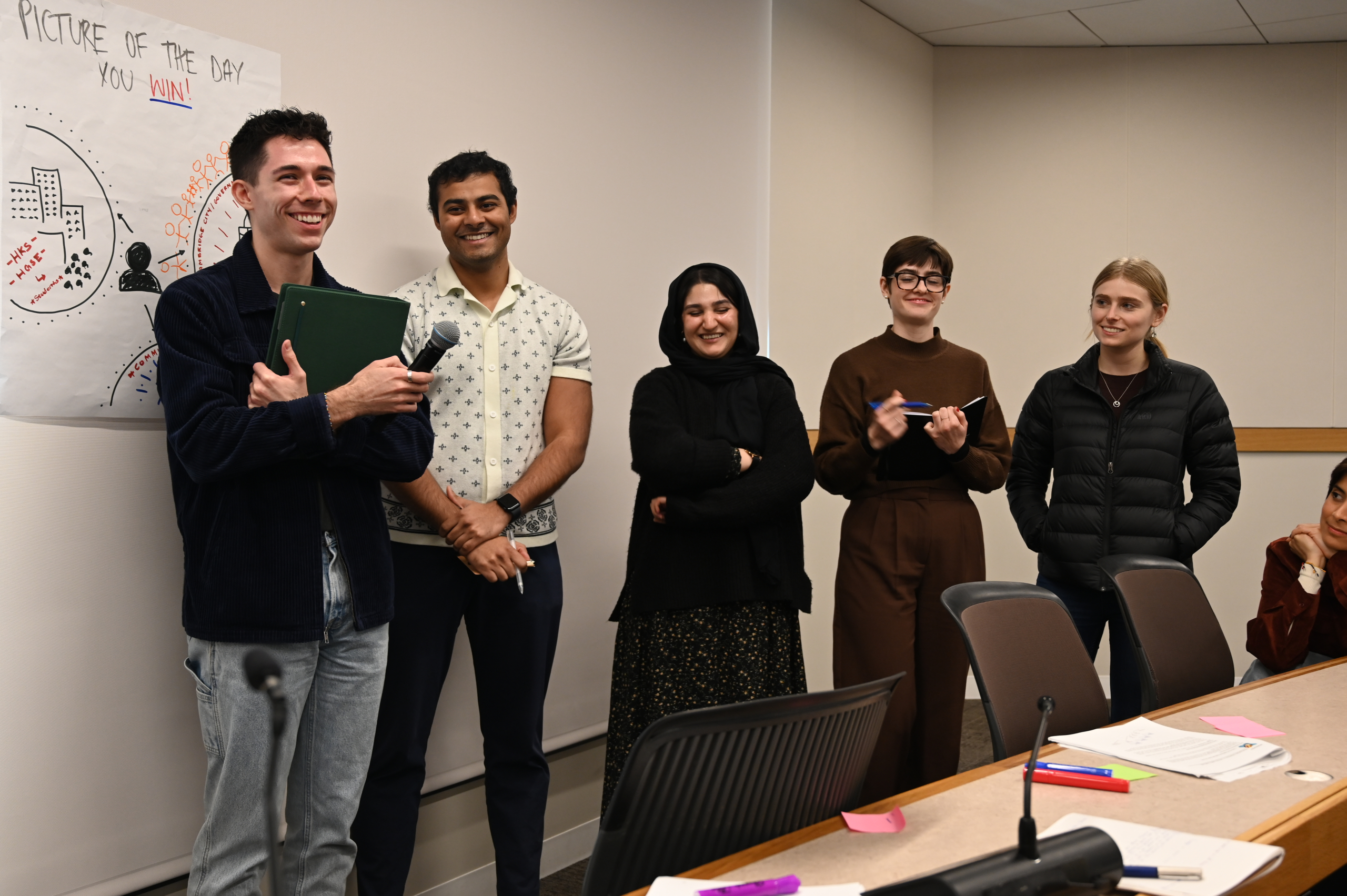 Five people stand facing the camera smiling and laughing, one more sits down on the right hand side. Zainab Azim stands in the middle.