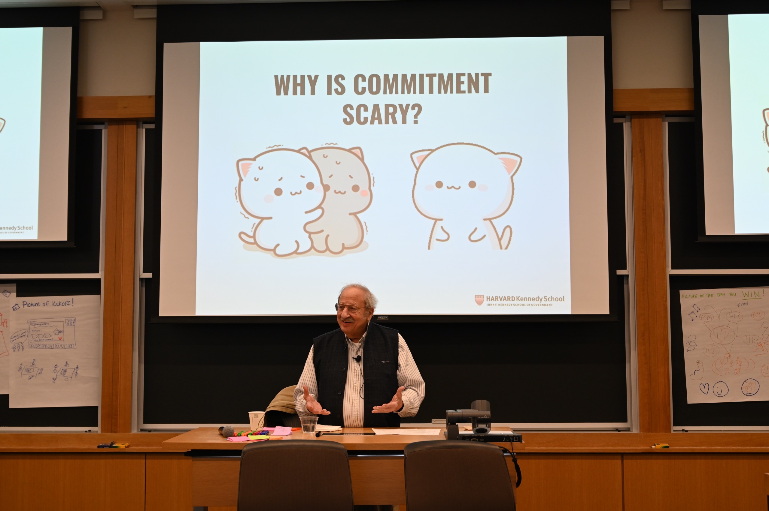 Marshall Ganz standing and smiling at the front of a Harvard lecture room, with a slide projected on the screen behind him. The slide says, 'Why is commitment scary?', and features an illustration of two cats shaking and hugging each other and another cat looking at them smiling.