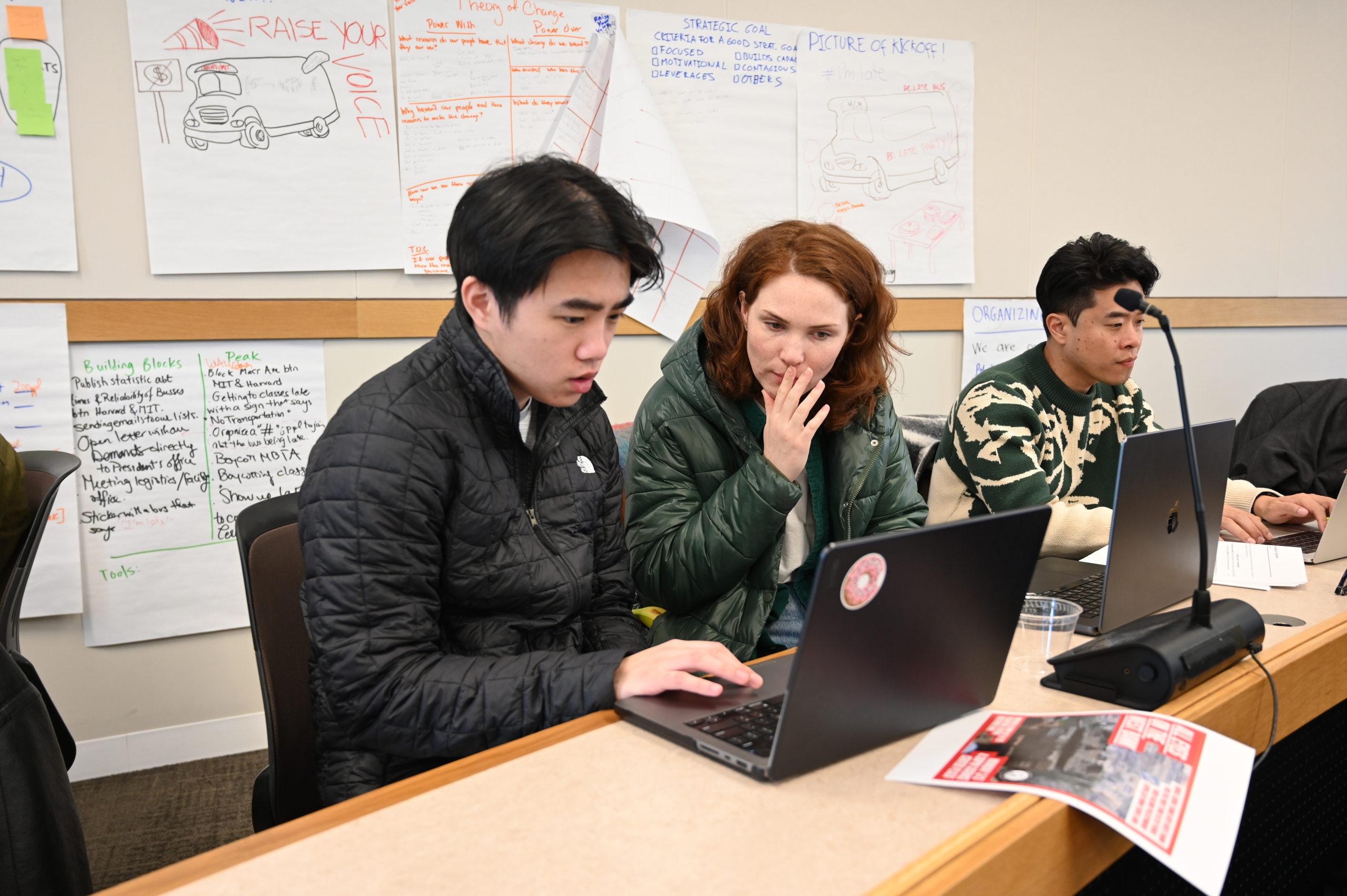 Three people sit at a desk working on computers. Maggie Hughes is sat in the middle, in discussion with the student on the left. Posters with hand-written text are pinned to the wall behind.