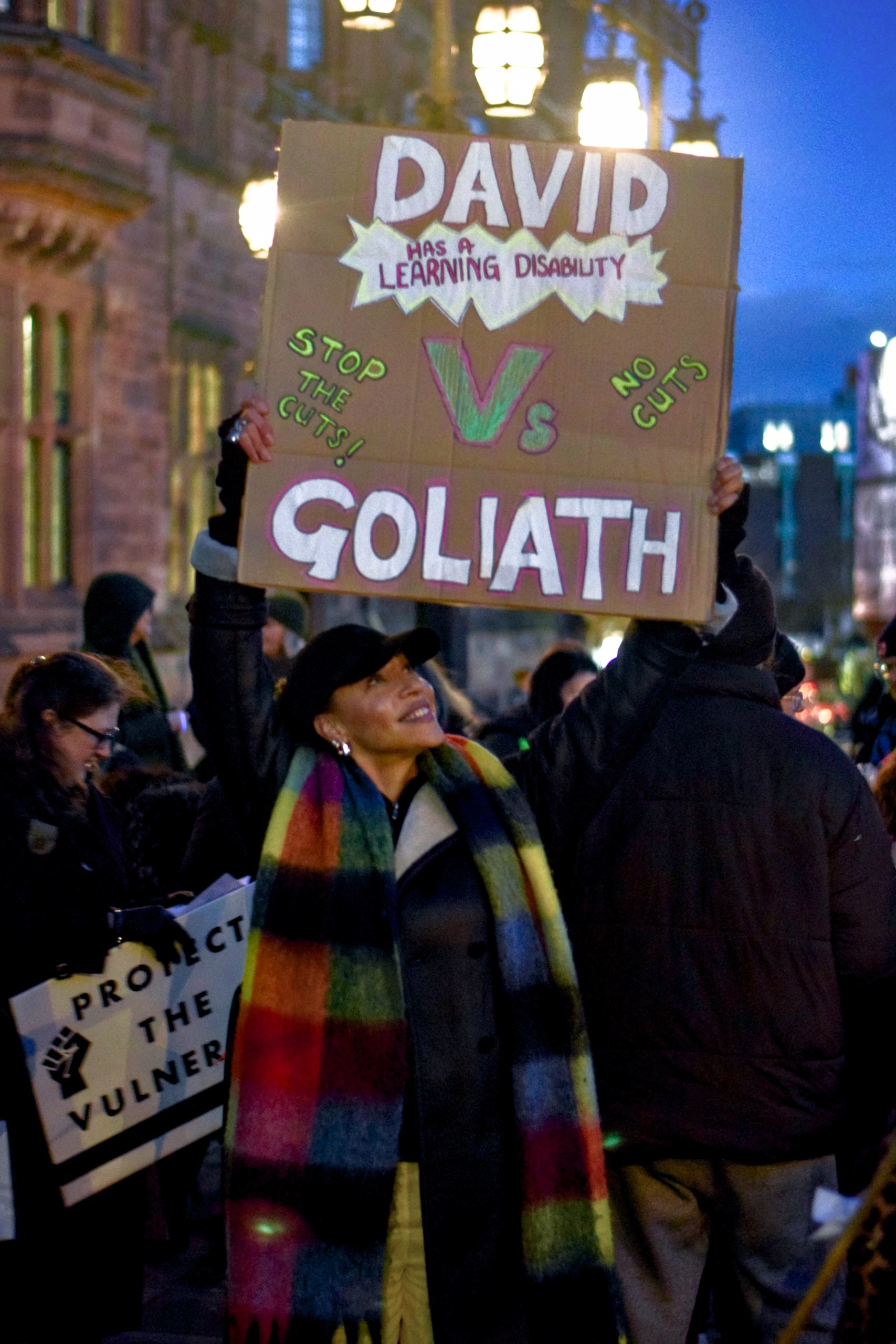 A person standing in the street amongst a crowd holding up a sign that says, 'David (has a learning disability) vs Goliath. Stop the cuts! No cuts.'
