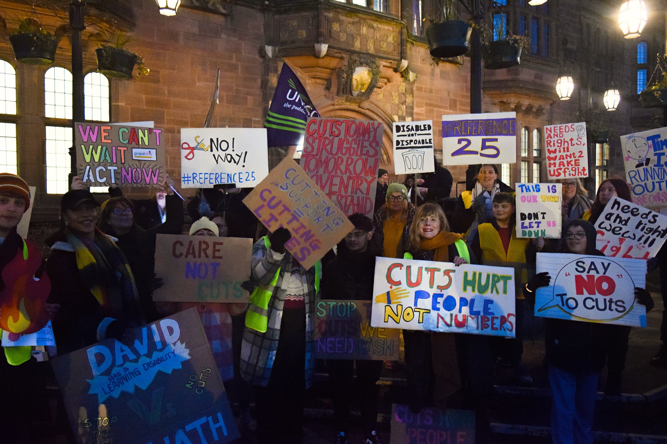A group of people protesting, lit by street lights. They are holding placards with various slogans against council funding cuts.