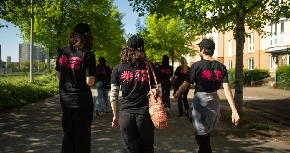 A group of canvassers walk down a residential street. There are houses to the right an a park to the left. Trees line both sides of the street. Three people wearing Green New Deal Rising t-shirts and black baseball caps are in the foreground, facing away from the camera. Three more people are in the background, waking the same direction but further ahead.