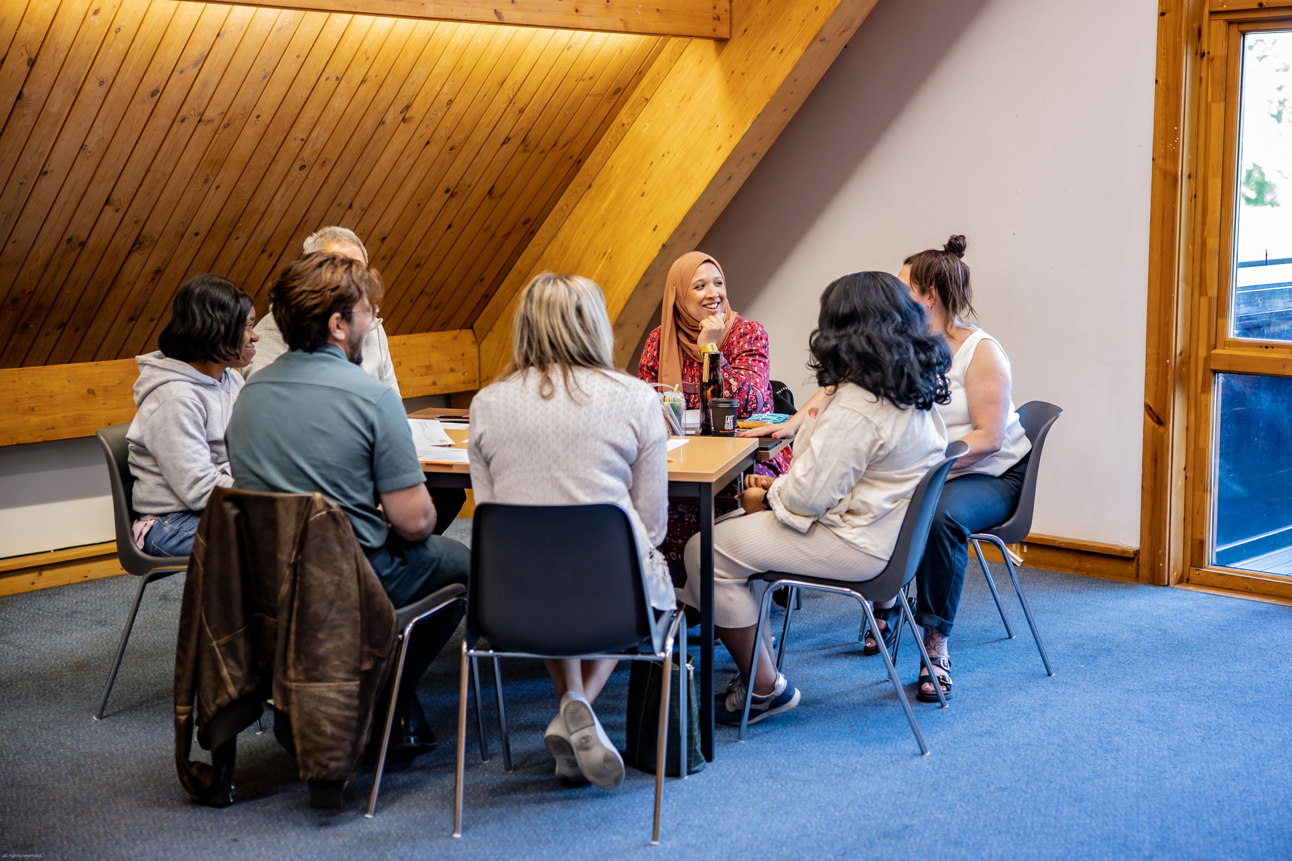 Seven people sit around a table with paper and pens. They are looking at the person at the right end of the table. The only person whose face is fully visible is a smiling woman in a hijab.