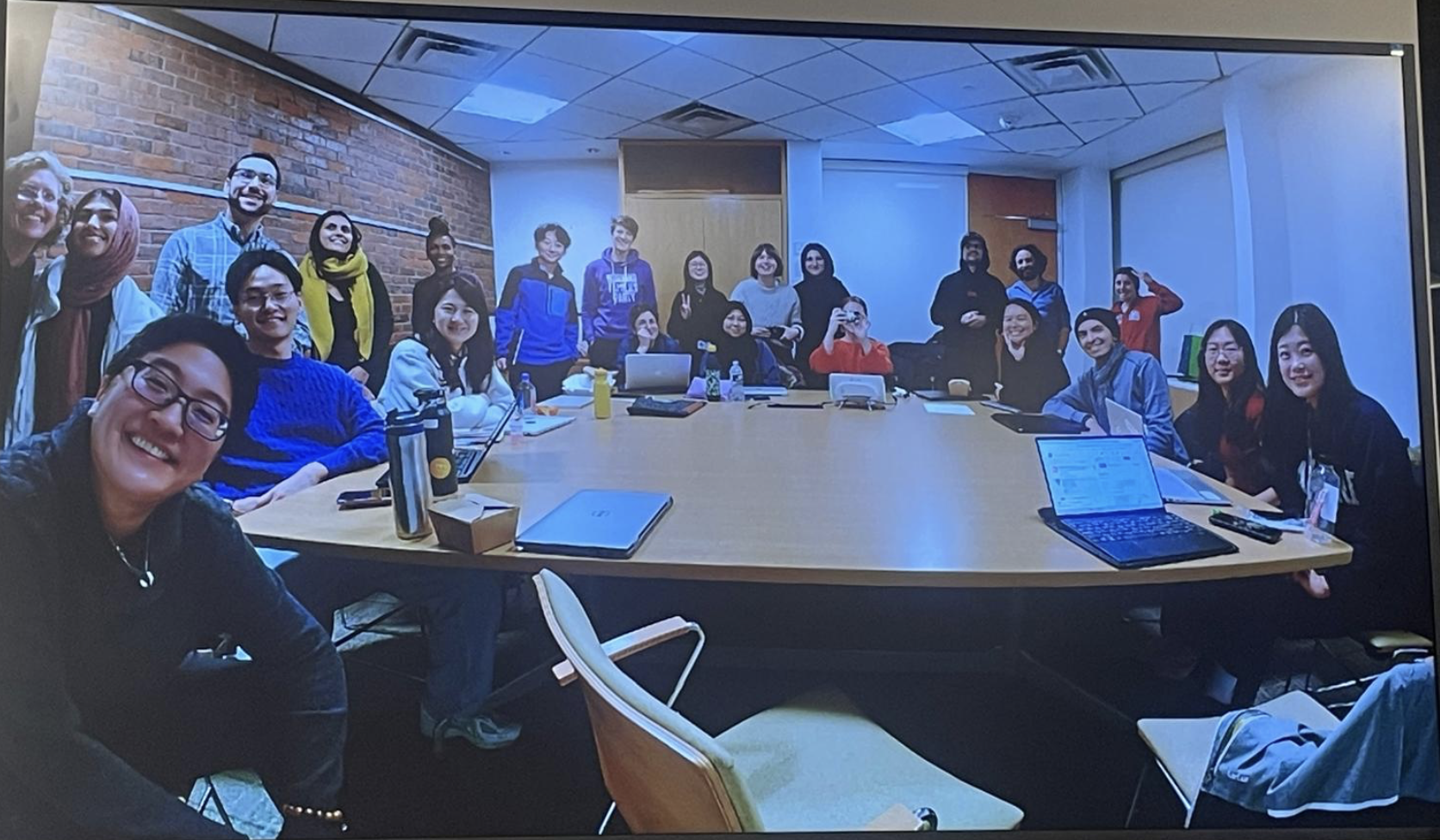 A diverse group of 23 people sit and stand around a large table, smiling at the camera.