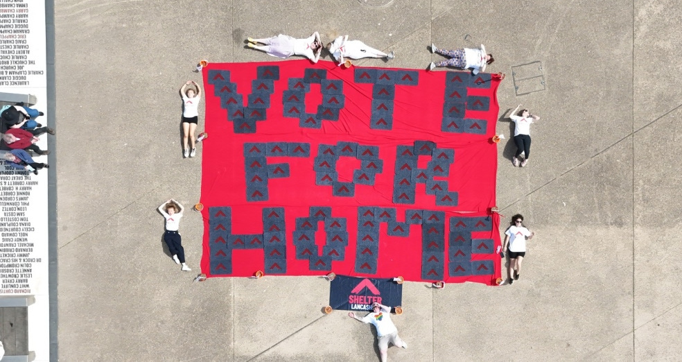 A birds eye view from high in the air showing a huge banner laid flat on concrete reading, 'Vote for home'. there are people lying on the floor around the banner.