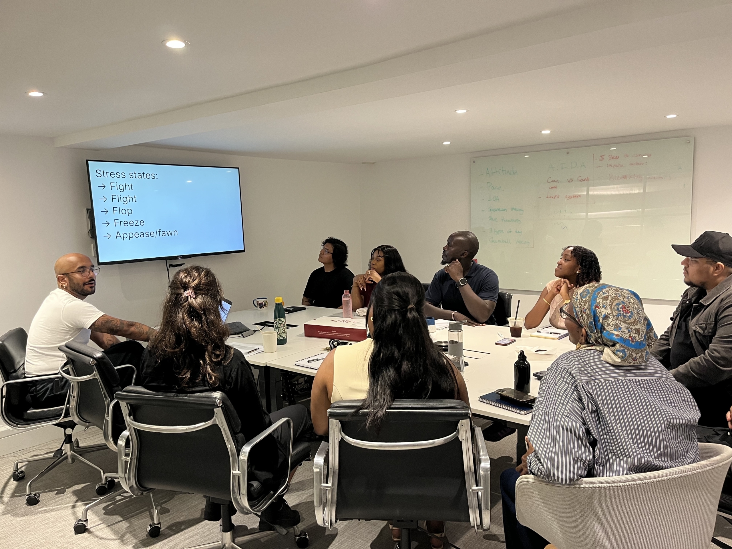 Eight people sit around a table looking at screen on the wall. Kennedy Walker sits on the left nearest the screen and is talking to the group. The screen reads: 'Stress states: Fight, Flight, Flop, Freeze, Appease/fawn.'