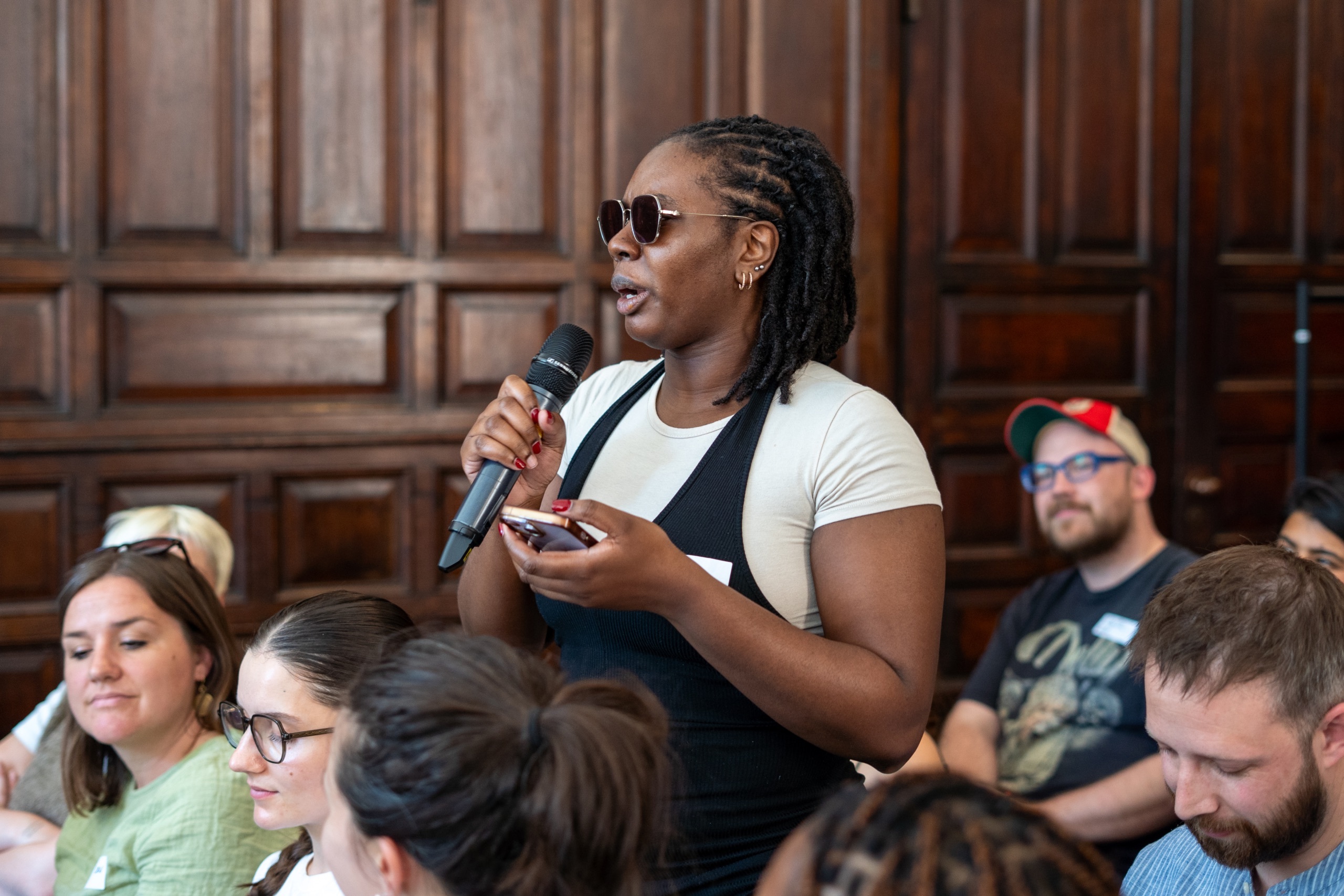 Jemmar, from Collective Punishment, is speaking into a microphone held in their hand. They are standing up amongst a seated audience.