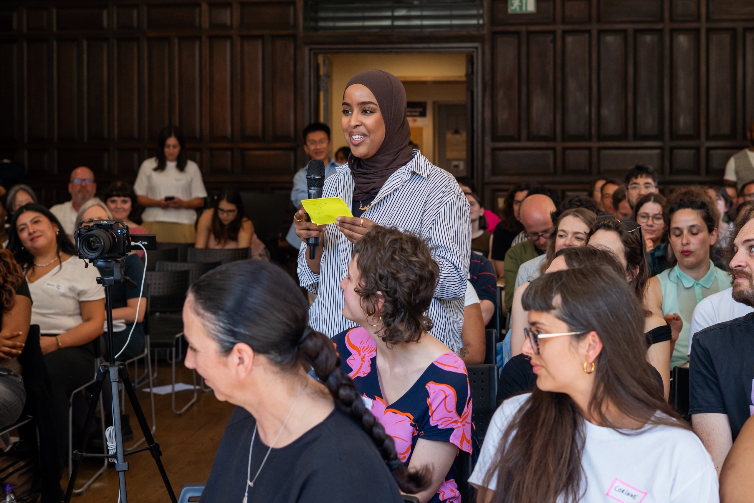 Fatima, from Green New Deal Rising, is speaking into a microphone held in their hand. They are standing up amongst a seated audience.