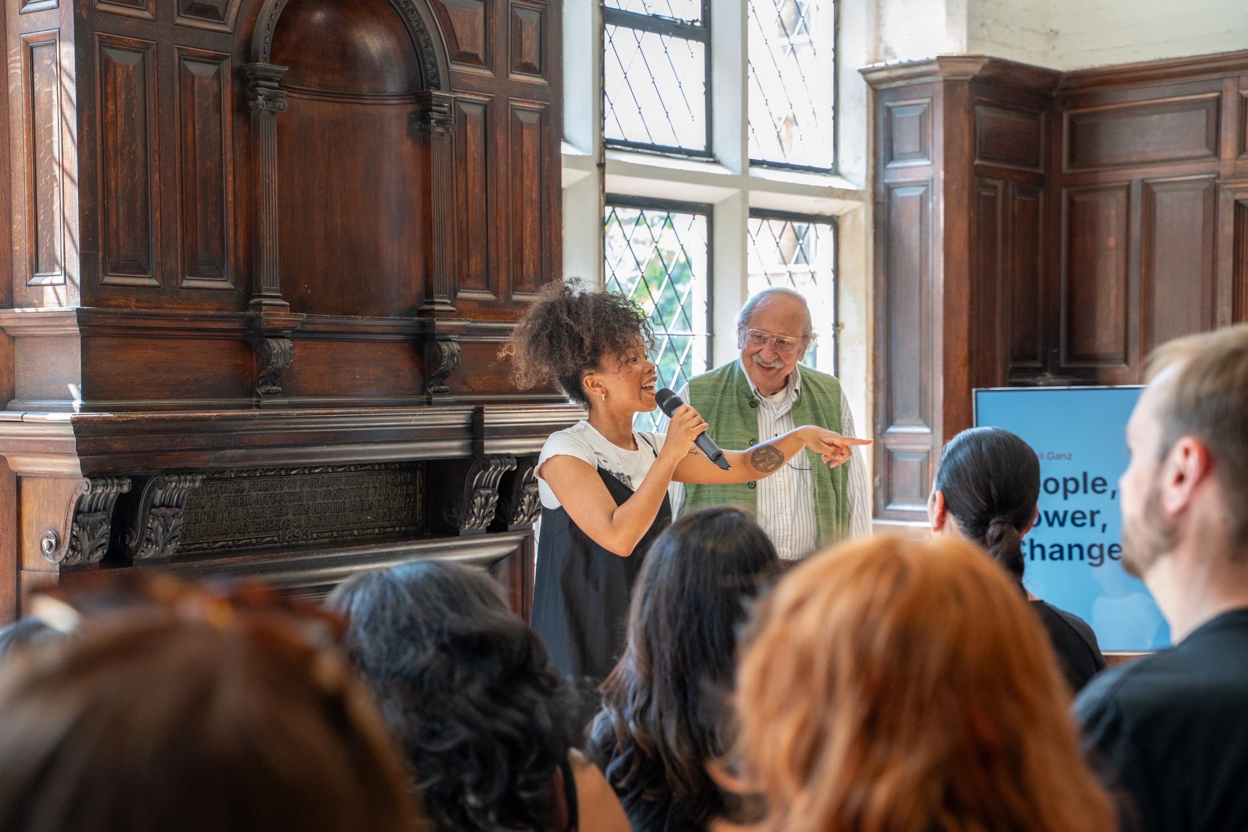 Amina Gichinga is stood on stage, alongside Marshall Ganz, in front of a standing audience. Amina is singing into a handheld microphone whilst pointing to the audience. Marshall is standing and smiling at Amina.
