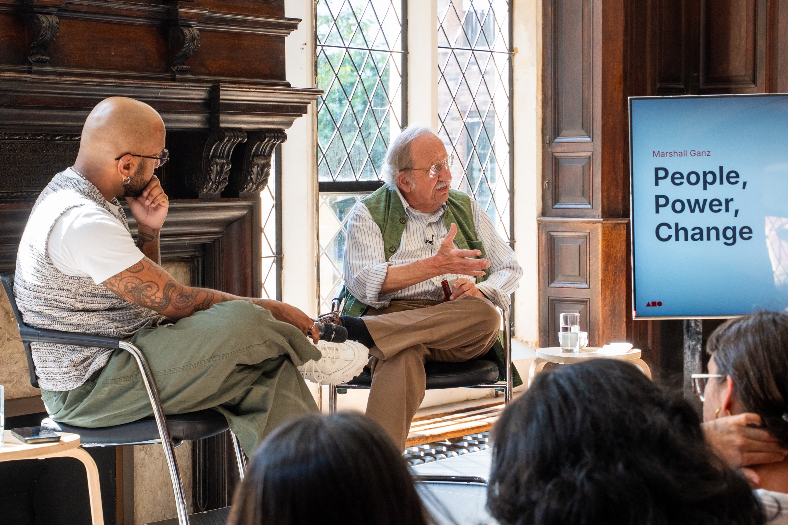 Kennedy Walker and Marshall Ganz are sat on chairs on a stage in front of an audience. Marshall Ganz is talking and gesturing towards the audience.