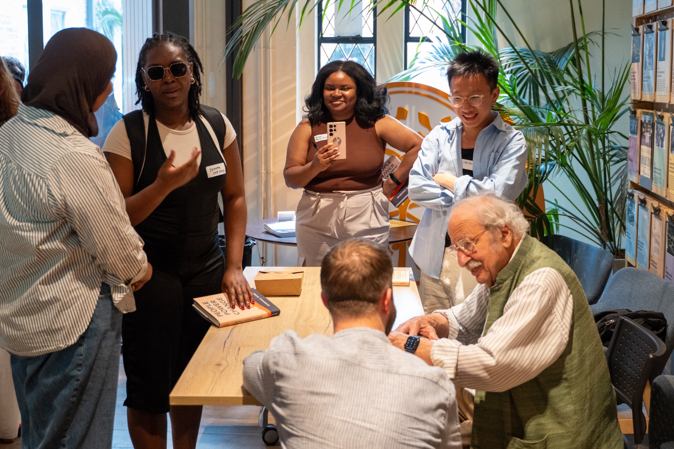 Marshall Ganz sitting at a table signing a book and talking to someone crouched next to him. There are other people standing around chatting, taking pictures, and waiting to get their books signed.