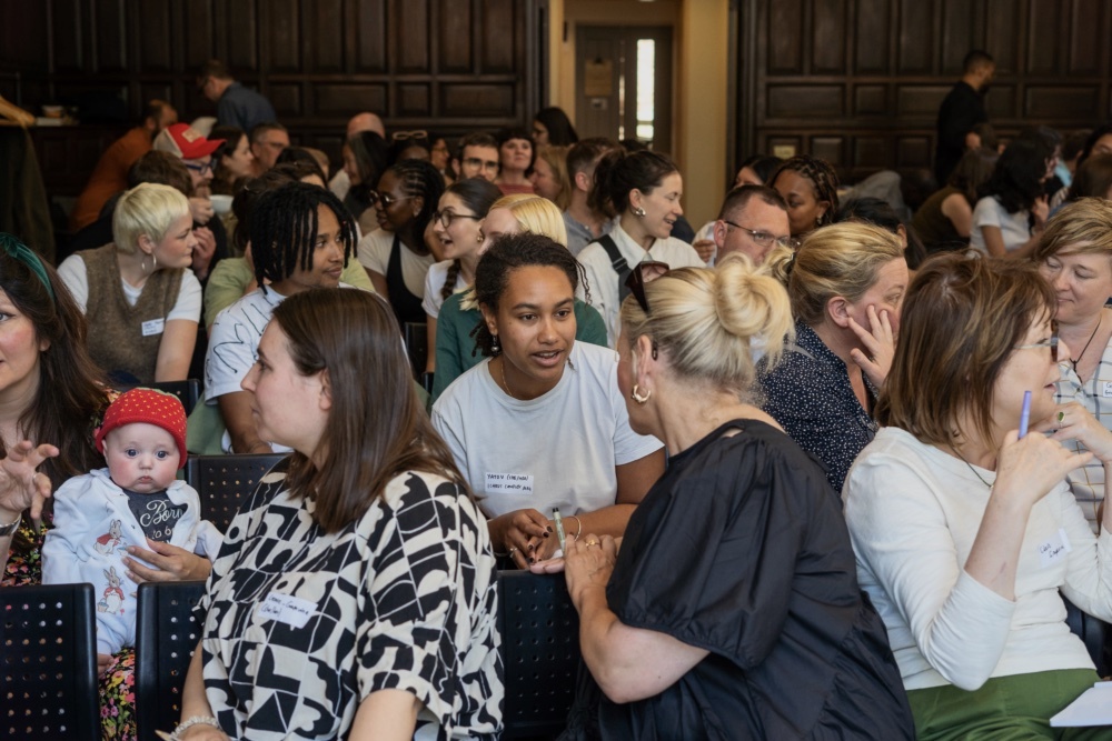 A picture looking back at the audience from the front of the room. The audience are sat down in rows of chairs and are engaged in conversation with people next to them and behind them.