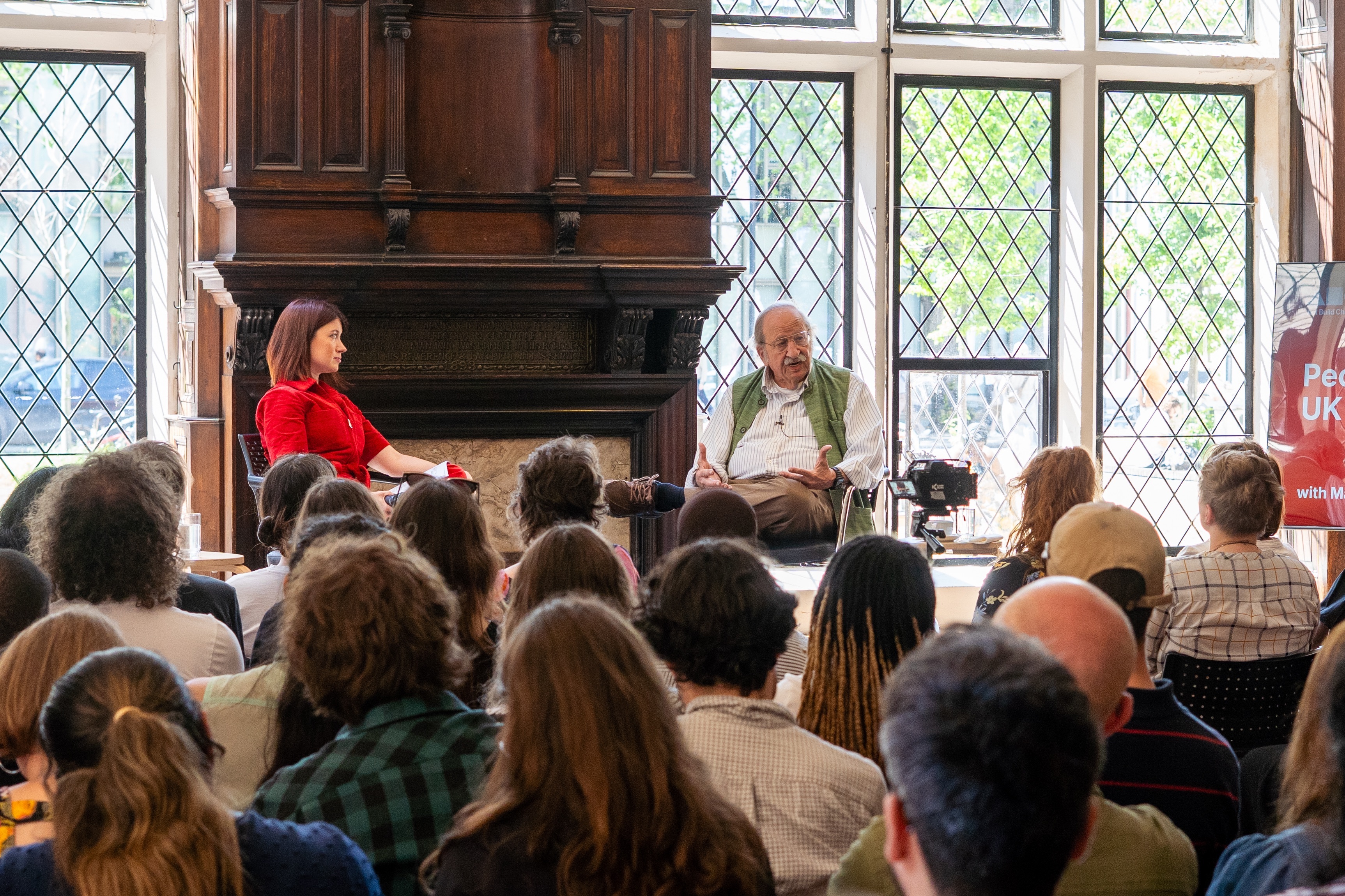 Stephanie Wong and Marshall Ganz are sat on chairs on a stage in front of an audience. Marshall Ganz is talking and gesturing towards the audience.