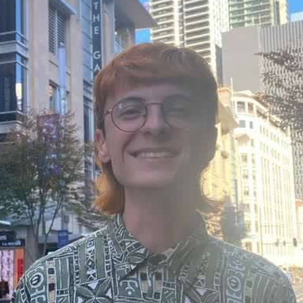 A picture of Billy Wassell. They are a white non-binary person which ginger hair and glasses. They are wearing a green patterned shirt.