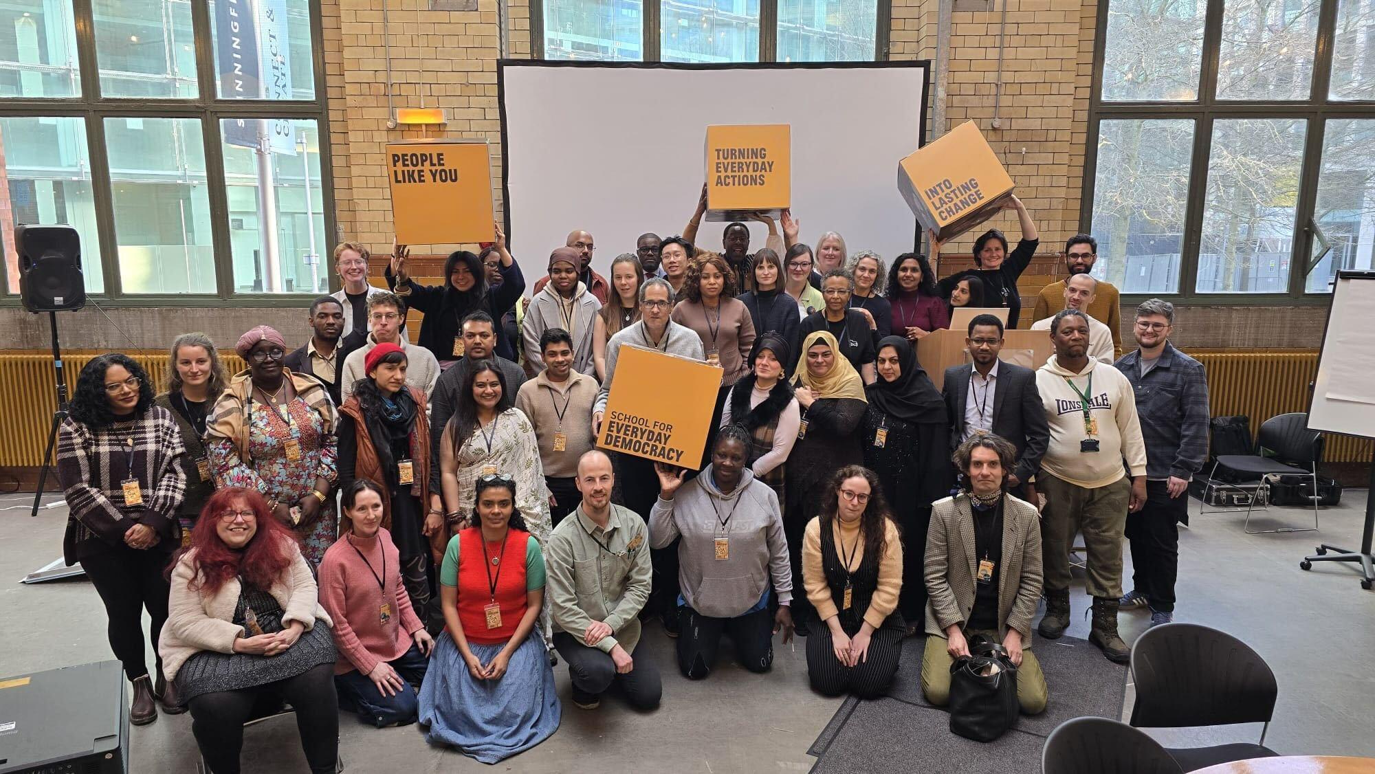 A group of 35 people smile at the camera. The people in the group are a range of ages and from a range of different countries. Some of them hold signs, these read: 'People Like You' 'Turning everyday actions into lasting change' and 'School for Everyday Democracy'