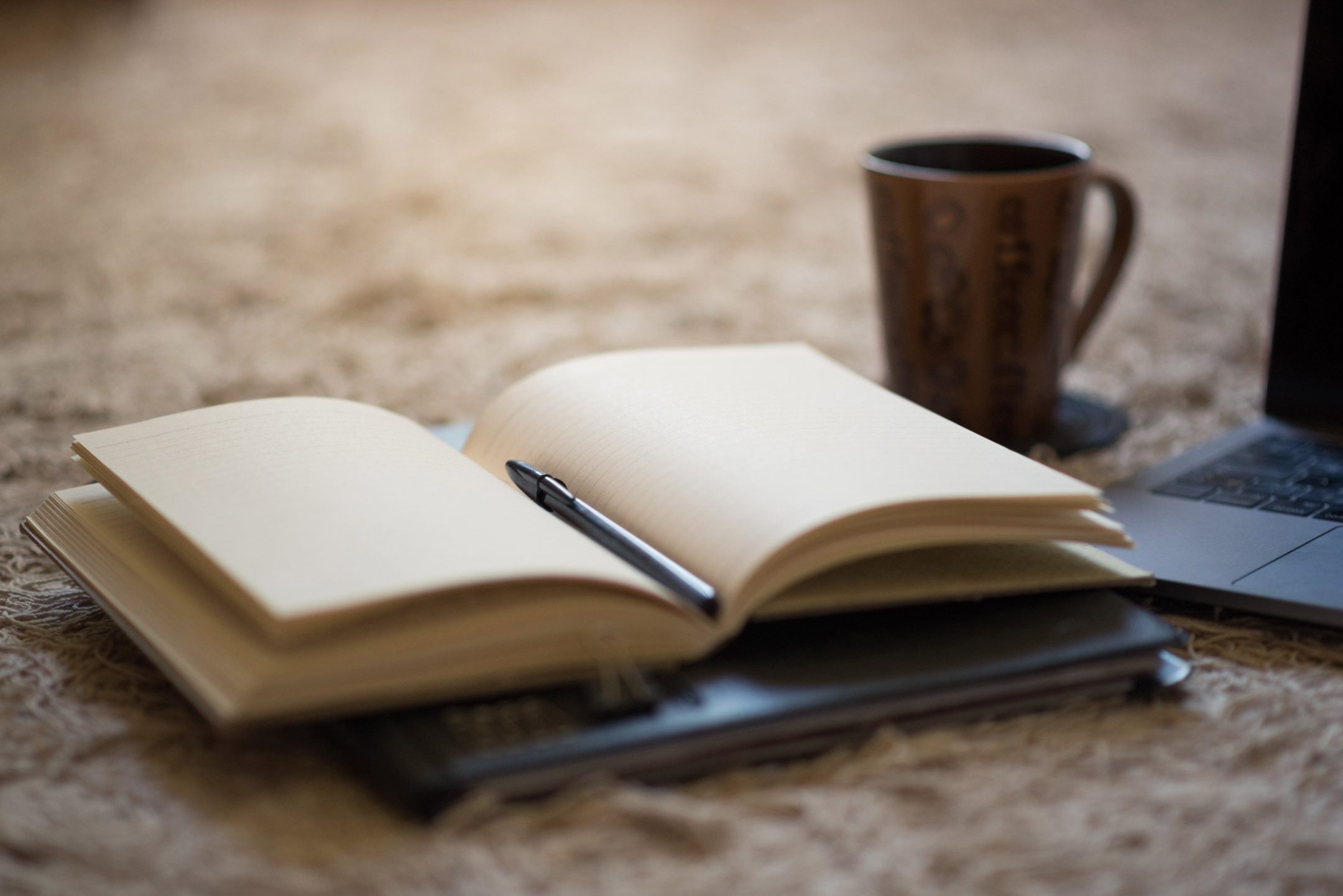 An open notebook with a pen in the fold, and a drinking mug and laptop in the background.