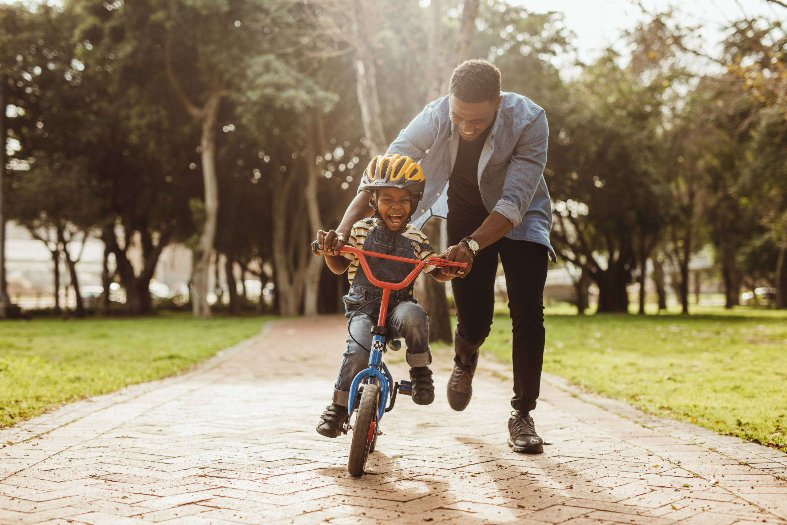 An adult pushing a young child on a bike, appearing to be helping them learn to ride a bike. The child looks happy and apprehensive at the same time.