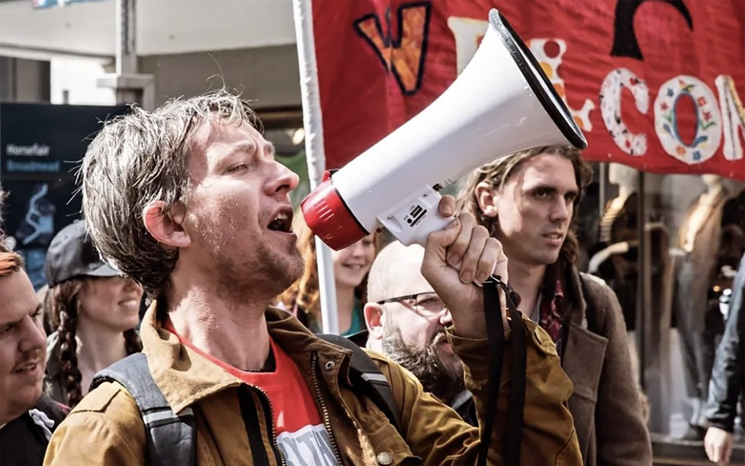 Louie Herbert, a community organiser, shouting through a megaphone at a march, standing in front of other marchers holding placards.