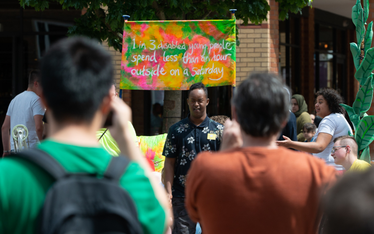 An image of a young woman telling her story to a crowd of people with a colourful banner behind her.