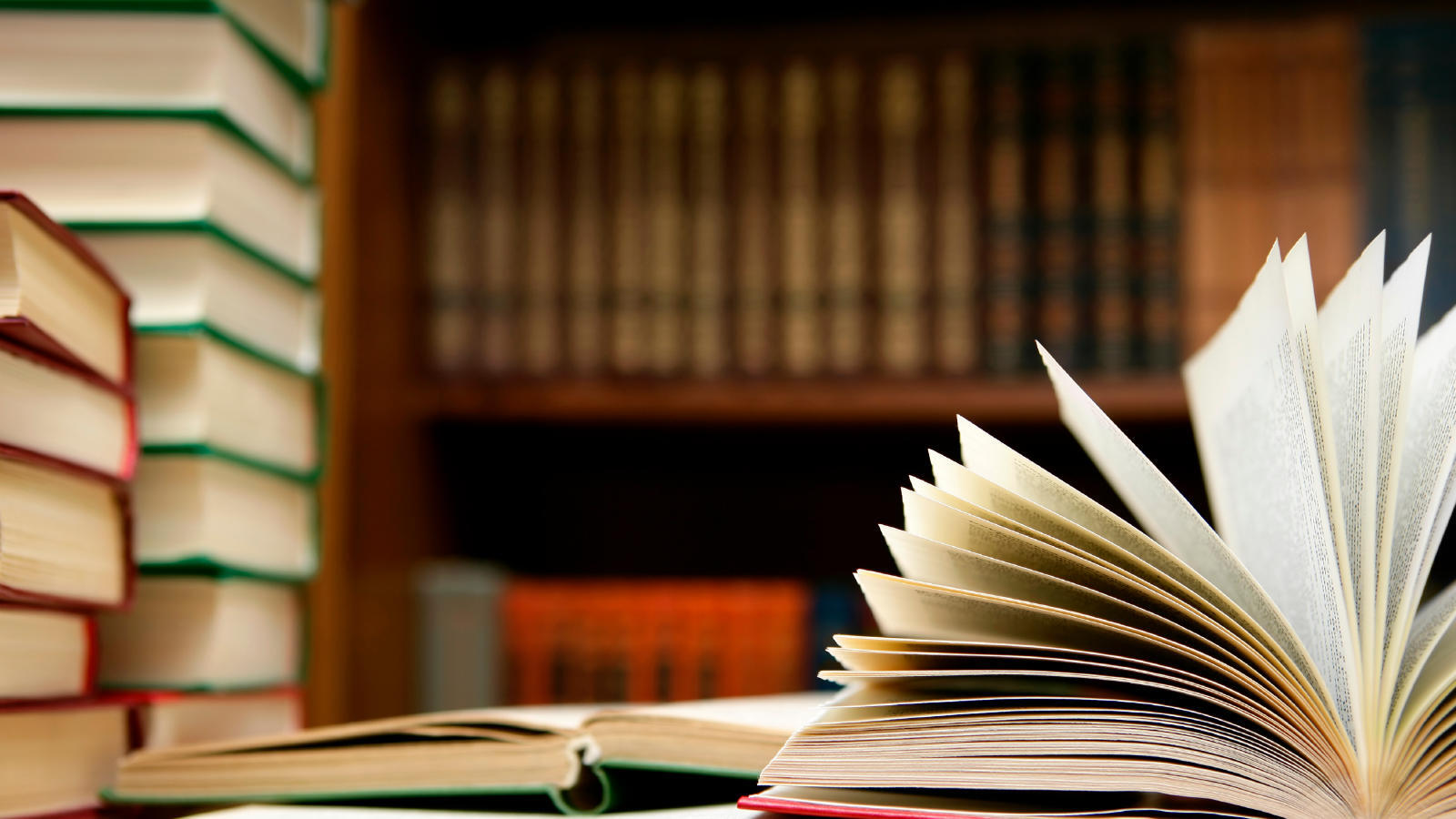 An image of an open book next to a stack of books with a book shelf in the background.