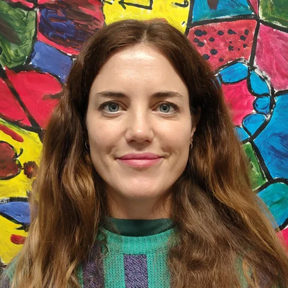 Lou is a white person with light blue eyes and long brown hair. They stand in front of a colourful painted wall.